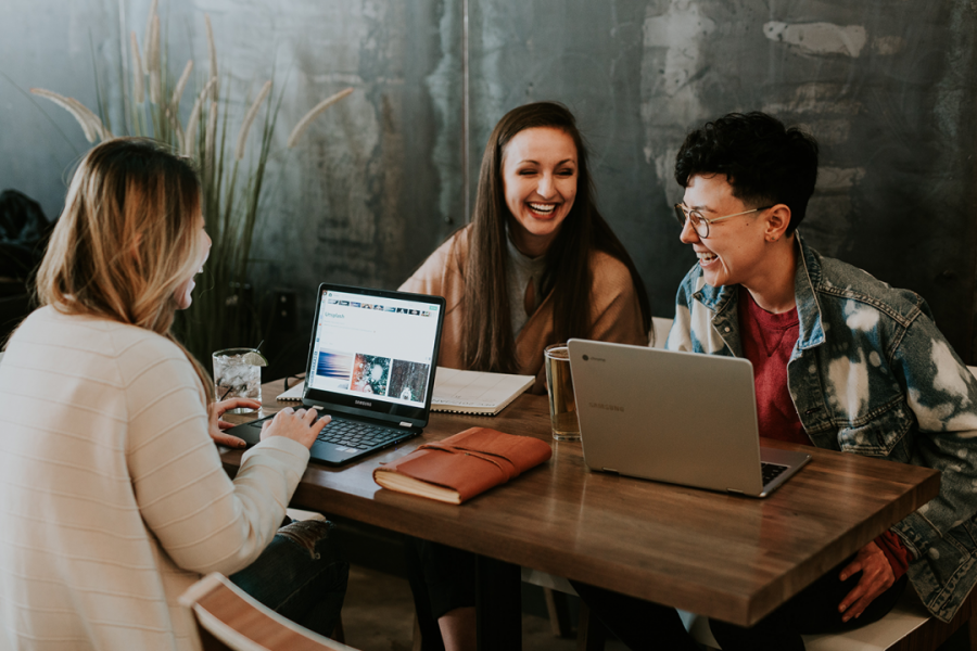Three people sitting at a wooden table using laptops and laughing